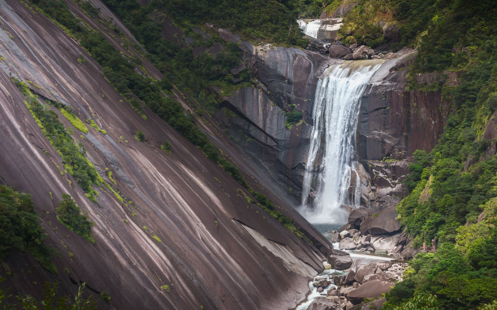 A waterfall pouring into a rocky gorge beside a vast smooth granite face