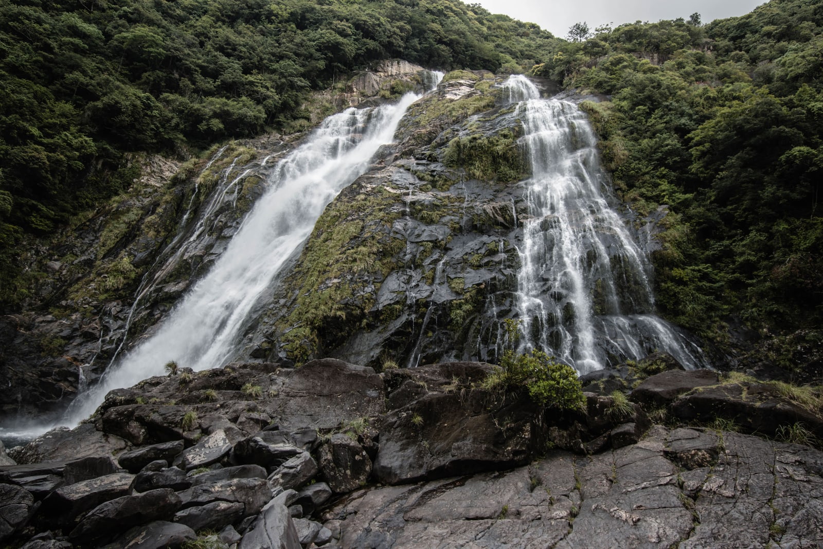 A tall waterfall falling in two streams over dark rock, seen from wet boulders at its foot on Yakushima