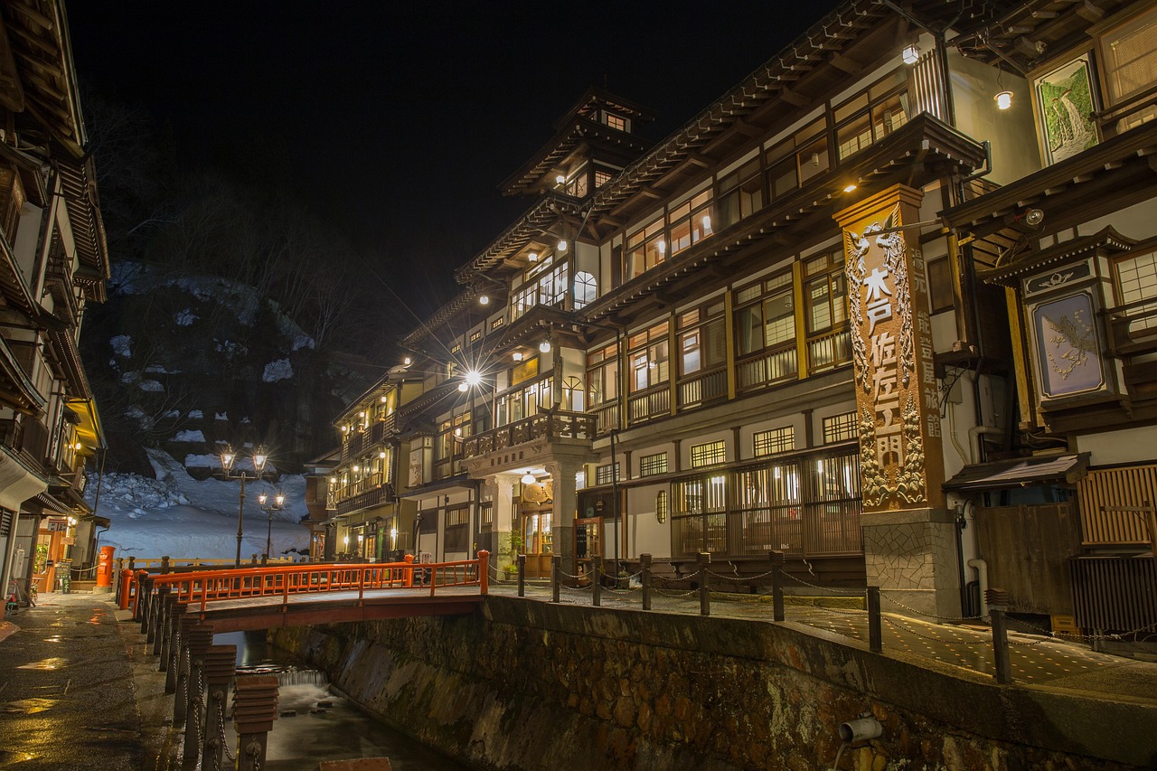 Wooden inns lit at night along a river at Ginzan Onsen, a red-railed footbridge and snow on the slope behind