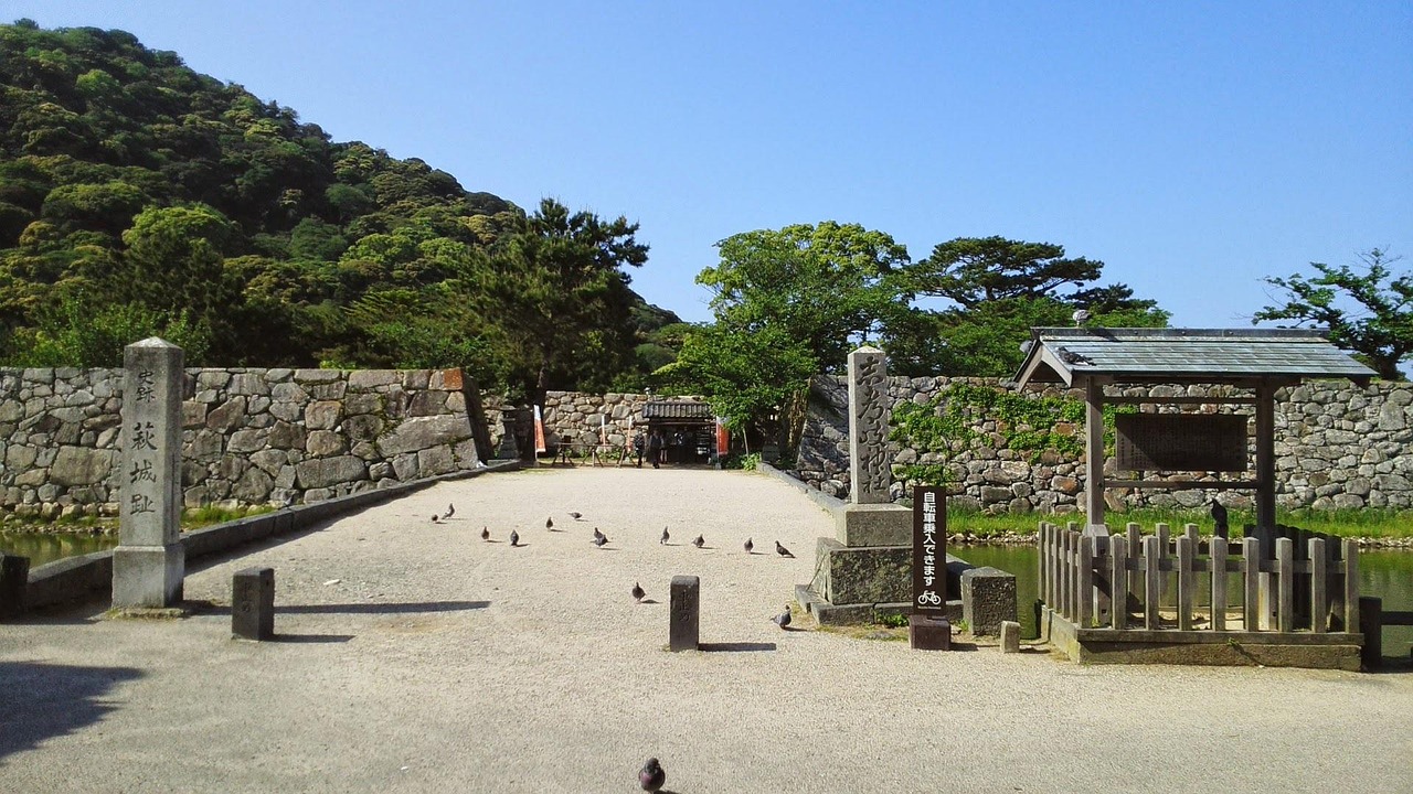 The entrance to the Hagi Castle ruins: a gravel approach between stone walls, a carved stone marker, a wooded hill behind