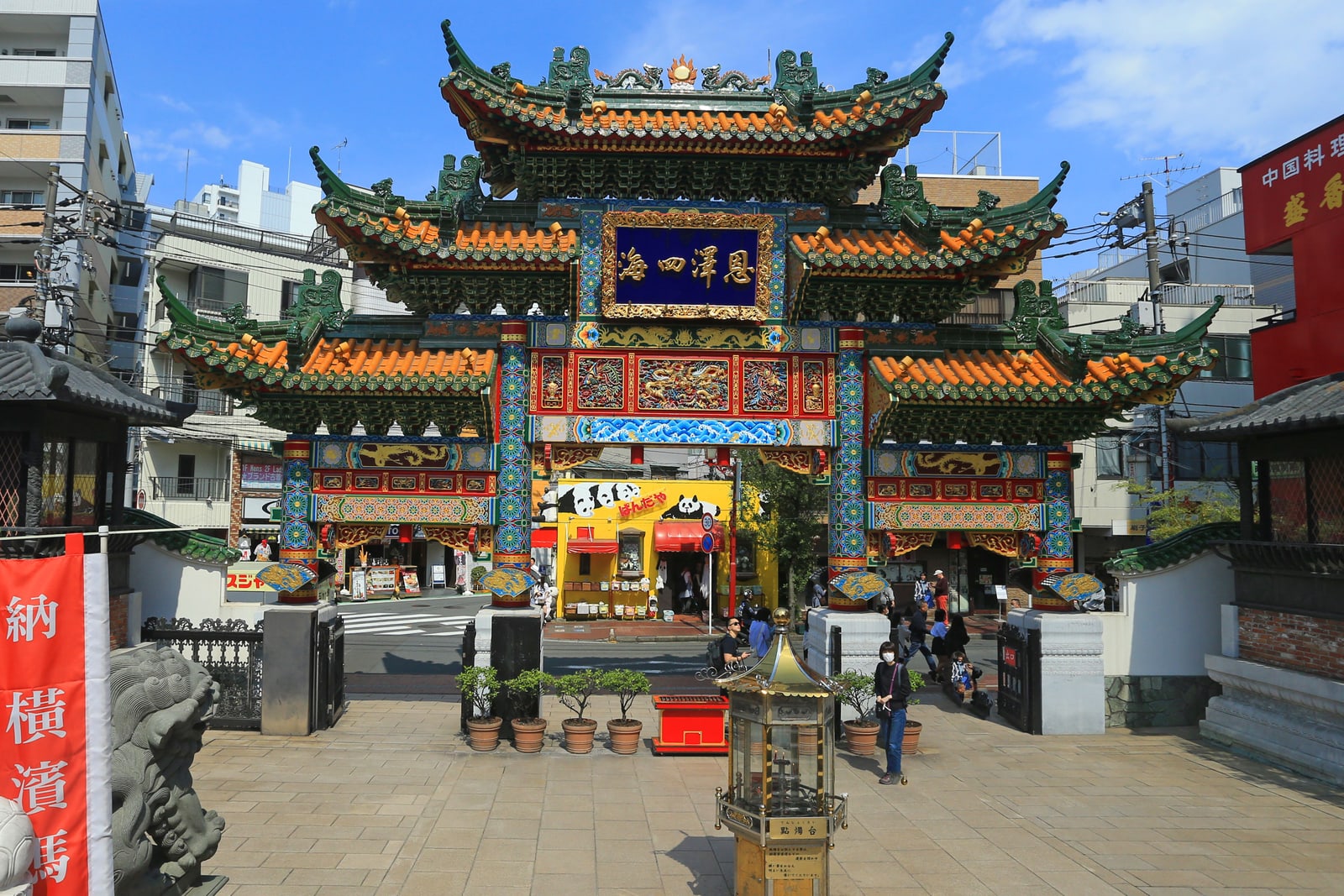 A brightly painted multi-roofed gate at Yokohama Chinatown with a blue plaque of gold characters, a stone lion and a few pedestrians in front