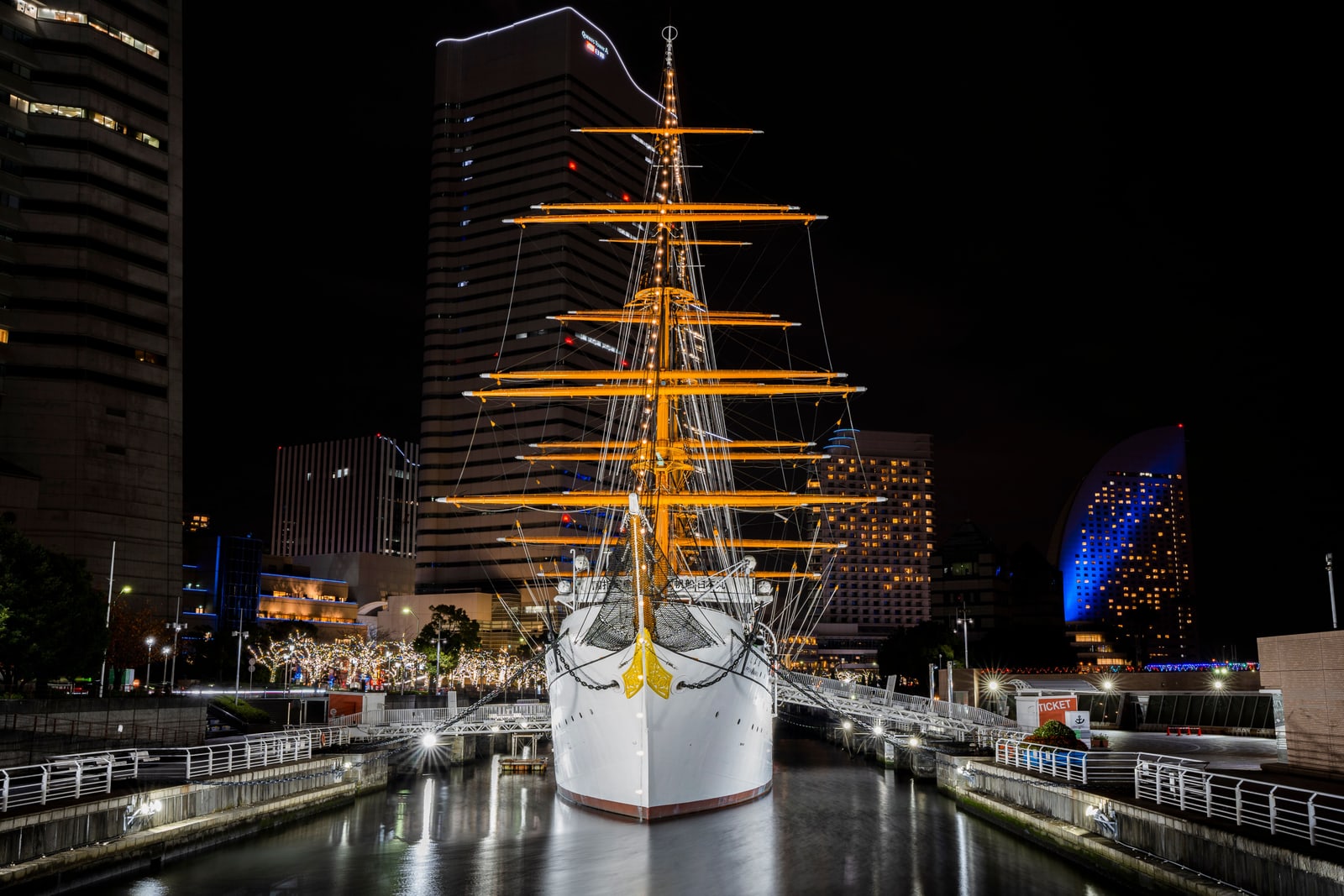 The white bow and lit yards of the sailing ship Nippon Maru in its dock at night, Minato Mirai towers behind