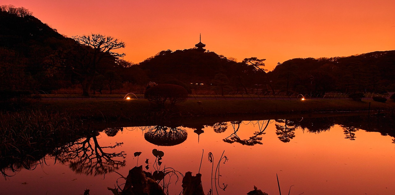 A three-storied pagoda silhouetted on a wooded hill over a still pond at dusk, the sky orange
