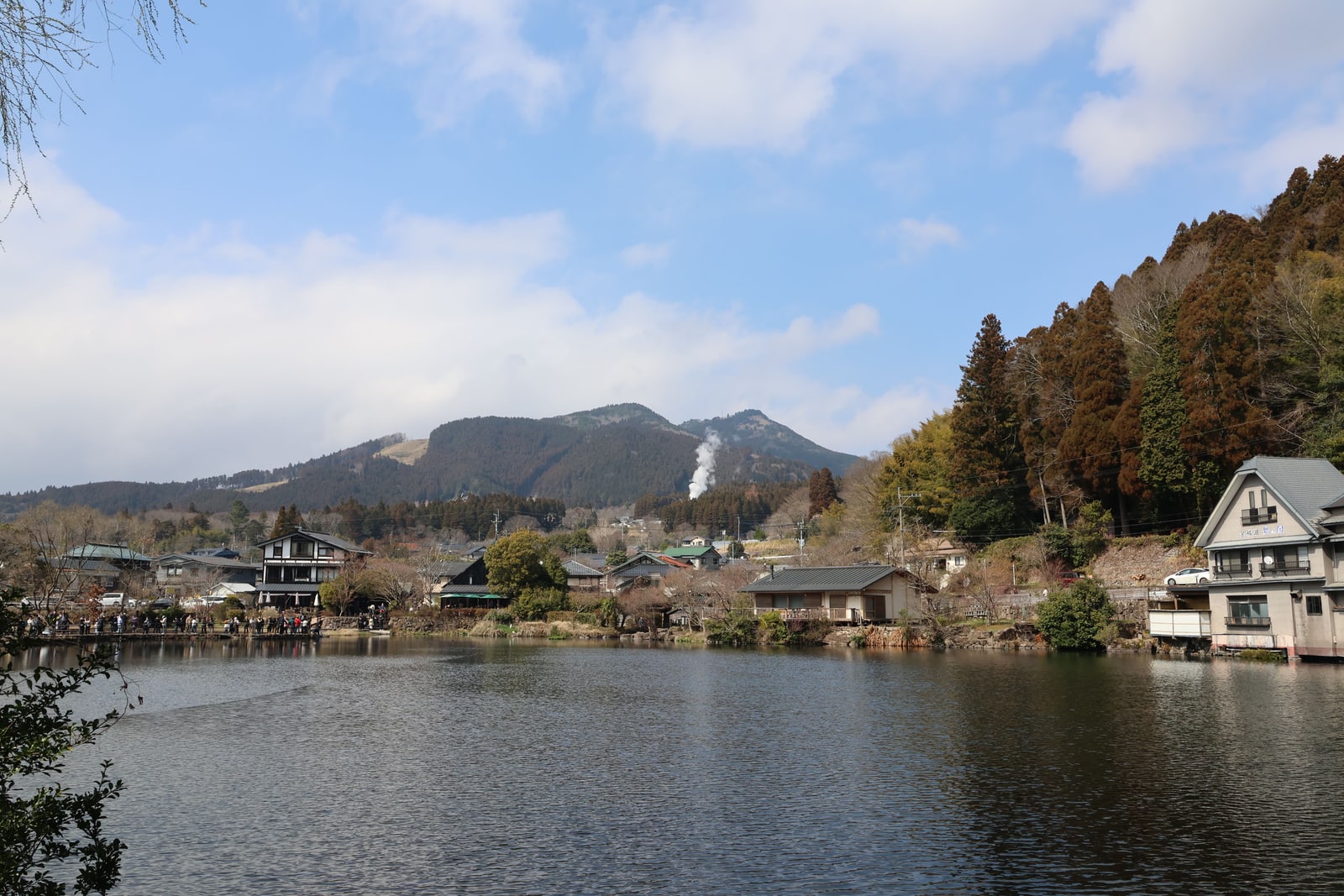 A lake with houses on the far shore and white steam rising from the wooded hillside behind