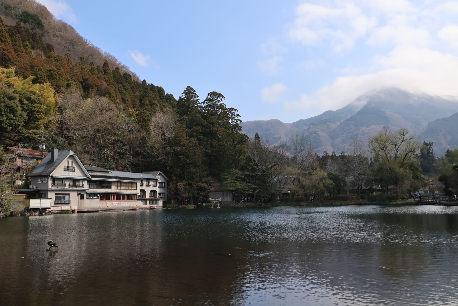 A small lake below a wooded hill, with a building at the water's edge, a small torii on the shore and a cloud-capped mountain